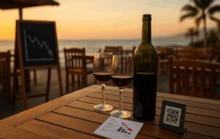 Botella y dos copas de vino en terraza frente al mar; pizarra con gráfica descendente y mesas vacías al atardecer en el sur de Tenerife.